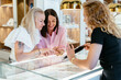 Jewelry boutique owner or shop assistant showing tablet with assortment on website on Internet, helping two women friends trying on ring at modern interior of jewelry shop.