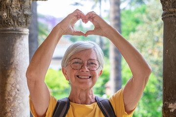 Sticker - Happy senior woman tourist traveling in Seville makes heart shape in the Alcazar park looking at camera enjoying vacations