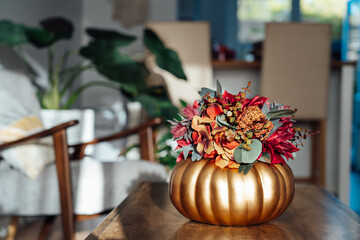Autumn bouquet of bright artificial flowers in a golden pumpkin vase on the wooden coffee table with modern living room interior background. Hygge home fall decor for a cozy home atmosphere.