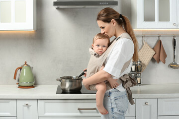 Wall Mural - Mother holding her child in sling (baby carrier) while cooking in kitchen