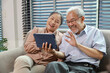© feeling lucky - Happy smiling asian senior couple sitting on sofa and using tablet while online video call with friend or relative cousin at home living room. Internet information technology and lifestyle concept.