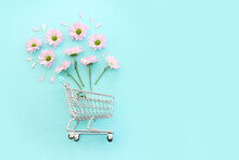 Shopping Cart With Flowers Over Pastel Blue Background. Holidays Shopping And Sale Concept