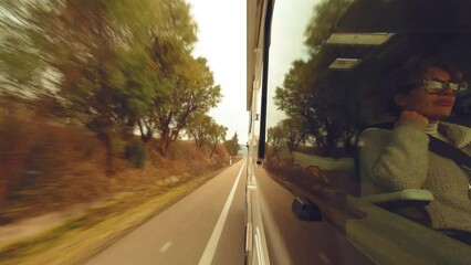 Wall Mural - Outside window view of woman passenger sitting inside a camper van vehicle traveling on a long straight road with sunset golden hour reflection on glass. Tourism and transport. Holiday vacation life