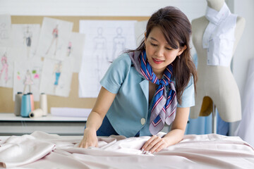 Young asian dressmaker in striped scarf spread out a piece of fine beige fabric on workbench preparing to create cloth pattern. Fashion designer room.
