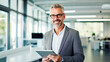 © BlazingDesigns - Portrait of mature man in office with tablet, smile and research for business website, online report or social media. Smiling mature corporate manager standing at office, looking at camera.