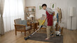 full length with side view asian man housekeeper in gloves and apron sprains his lower back and moans painfully while he is vacuuming floor in the living room