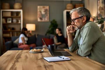 A senior man working on a laptop at home, while his family sitting on a couch.