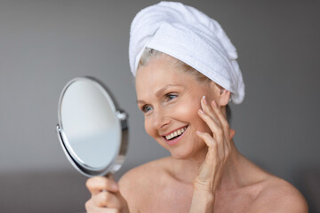 Wall Mural - Facial treatment concept. Beautiful middle aged woman in towel on head looking in mirror and touching her perfect skin