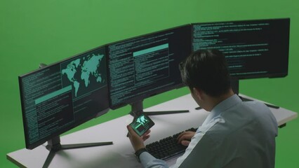 side view of Asian man developer working with mobile phone while write code with multiple computer screens in green screen studio
