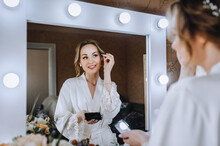 A Beautiful Smiling Bride Model Stands In The Morning In A Room, Indoors In White Lingerie In Front Of A Mirror With Lamps, Reflecting, Doing Makeup. Wedding Photography, Portrait.