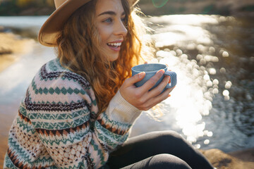 Beautiful woman traveler in a hat sits on a log near the river, drinks a hot drink from a thermos. Smiling female tourist enjoys the autumn landscape near the river. The concept of travel, relax.