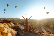 Woman Tourist In A Hat Stands With Her Back On A Mountain, Looking At The Balloons In Cappadocia, Turkey. Beautiful Woman On Top Of A Mountain Enjoys A Beautiful View. 