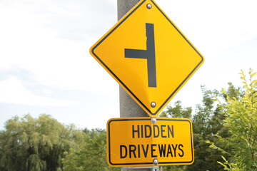 Sticker - hidden driveways rectangle sign with diamond sign above of sideways t to illustrate hidden driveways, yellow and black, trees and sky behind