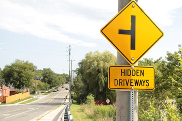 Sticker - hidden driveways rectangle sign with diamond sign above of sideways t to illustrate hidden driveways with bottom of bridge in background, yellow and black, trees and sky behind