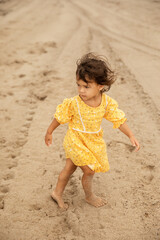 Little toddler girl in yellow dress walking on sandy beach, summer vacation concept