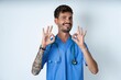 © Roquillo - young caucasian doctor man wearing blue medical uniform showing both hands with fingers in OK sign. Approval or recommending concept