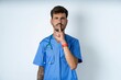 © Roquillo - young caucasian doctor man wearing blue medical uniform silence gesture keeps index finger to lips makes hush sign. Asks not to share secret