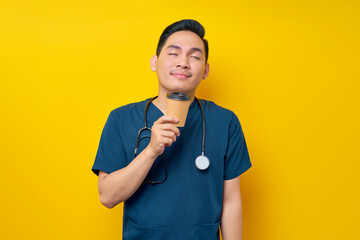 Handsome young Asian male doctor or nurse wearing blue uniform enjoying a drink of coffee isolated on yellow background. Healthcare medicine concept