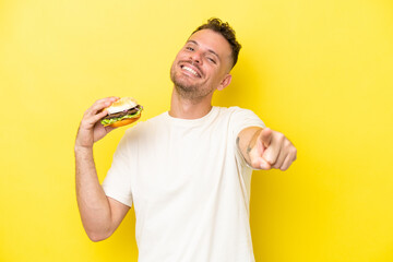 Wall Mural - Young caucasian man holding a burger isolated on yellow background pointing front with happy expression