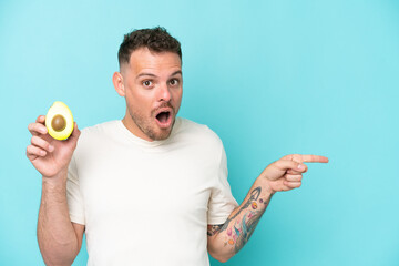 Wall Mural - Young caucasian man holding an avocado isolated on blue background surprised and pointing side