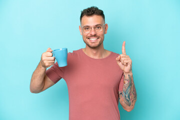 Wall Mural - Young caucasian man holding cup of coffee isolated on blue background pointing up a great idea