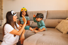 Family Doing Sleight Of Hand On The Sofa At Home, Laughing, With Funny Halloween Or Carnival Masks, Fun Time Of Your Kids With Their Mom