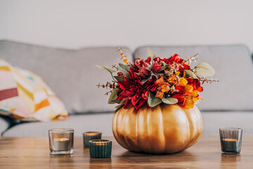 Autumn bouquet of bright artificial flowers in a golden pumpkin vase, burning candles on the wooden coffee table in living room interior with sofa. Hygge home fall decor for a cozy home atmosphere.