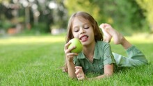 A Baby Girl Eats A Green Apple On A Green Lawn In Summer And Smiles Lying On The Grass, A Cheerful Child With An Apple, Children's Holidays, Close-up, Healthy Teeth