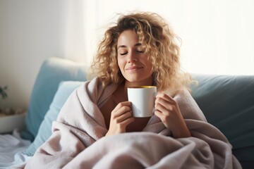 beautiful young blond dreamy woman sitting on bed wrapped in a blanket drinking tea or coffee