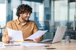 Successful hispanic financier on paperwork, young businessman investor checking papers, accounts contracts and statistical reports, man with laptop inside office smiling happy with achievement.