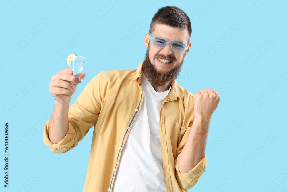 Cheerful young man with shot of tequila on blue background