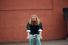 Long-haired Blond Bearded Man In Rain Jacket Riding A Bicycle Along The Street On Rainy Day Against Brown Wall Background 