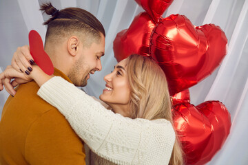 Young brunette man and blonde woman holding heart in her hands looking at each other and hugging with red foil ballons background. Happy couple in love. Care, tenderness and Valentines day concept.