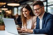 © amnaj - Cheerful businesspeople using a laptop in an office. Two happy young entrepreneurs smiling while working together in a modern workspace. Two young businesspeople sitting together at a table.