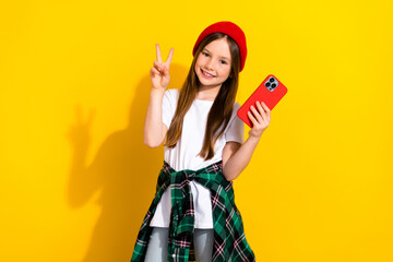 Poster - Photo portrait of cute little girl hold telephone showing v-sign wear trendy white clothes isolated on yellow color background