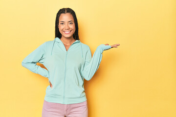 Indonesian sporty woman on yellow backdrop showing a copy space on a palm and holding another hand on waist.