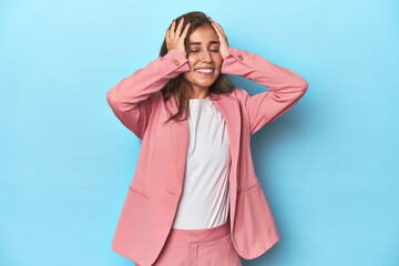 Wall Mural - Teen girl in chic pink suit on a blue background laughs joyfully keeping hands on head. Happiness concept.