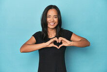 Filipina Young Woman On Blue Studio Smiling And Showing A Heart Shape With Hands.