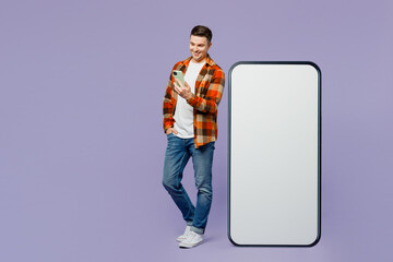 Wall Mural - Full body young man wears checkered shirt white t-shirt casual clothes stand near big huge blank screen mobile cell phone with area using smartphone isolated on plain pastel light purple background.
