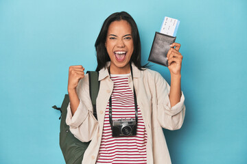 Filipina with camera, tickets, backpack on blue raising fist after a victory, winner concept.