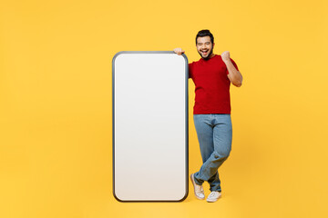 Wall Mural - Full body young Indian man wear red t-shirt casual clothes big huge blank screen mobile cell phone smartphone with area do winner gesture isolated on plain yellow orange background. Lifestyle concept