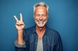 © Markus Schröder - Lifestyle portrait photography of a grinning mature man making a peace and love gesture with the fingers against a soft blue background. With generative AI technology