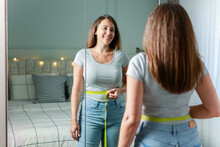 Smiling Young Woman After Weight Loss Measuring Waist In Front Of Mirror