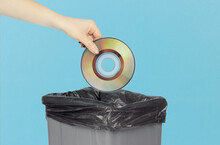 Throw Compact Discs Into The Trash Can, Compact Discs In Hand In Front Of The Trash Can,a Gray Trash Can On A Blue Background. Old Technology Concept