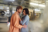 Fototapeta  - cheerful redhead man hugging young asian girlfriend in blurred public laundry
