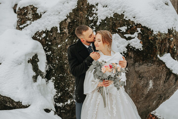 Wall Mural - Wedding in winter. Stylish bride in white poncho and groom in black coat in forest with white snow in cold weather. The groom kisses the bride against the background of snowy rocks