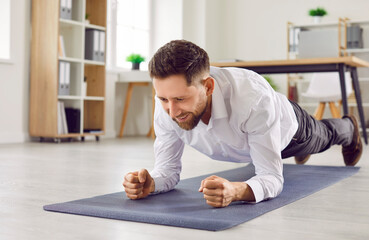 Portrait of a young cheerful business man doing plank exercise on yoga mat on the floor in office. Happy smiling company employee during the break. Workplace sport and fitness concept.