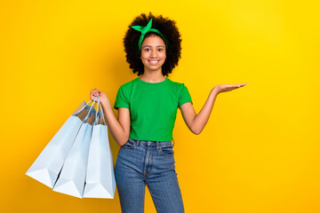 Poster - Portrait of beautiful teen girl hold boutique bags arm palm demonstrate empty space isolated on yellow color background