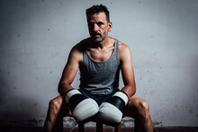 Serious Adult Man In Boxing Gloves Sitting On Wooden Stool In Room