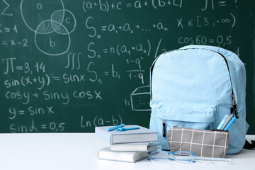 Blue school backpack with eyeglasses and stationery on white table near green chalkboard